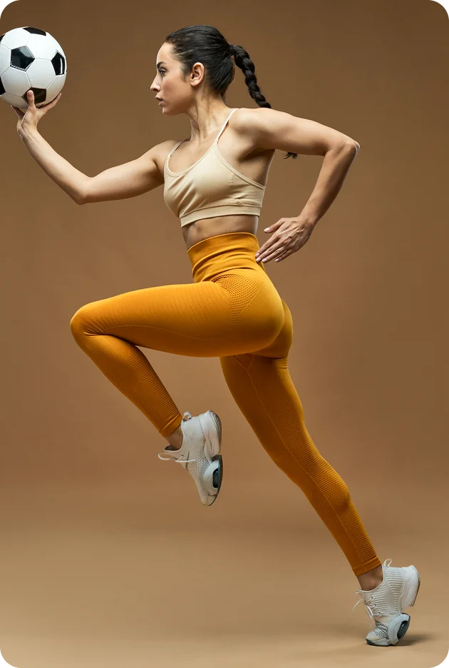 Athletic woman in mustard yellow high-waisted compression leggings and nude sports bra holding a soccer ball, jumping in dynamic pose.