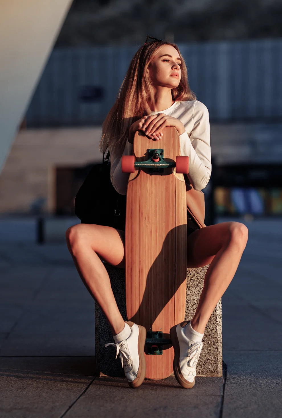 Lifestyle fashion banner featuring a young woman in white long-sleeve top and sneakers, sitting with a wooden longboard in an urban setting at sunset.