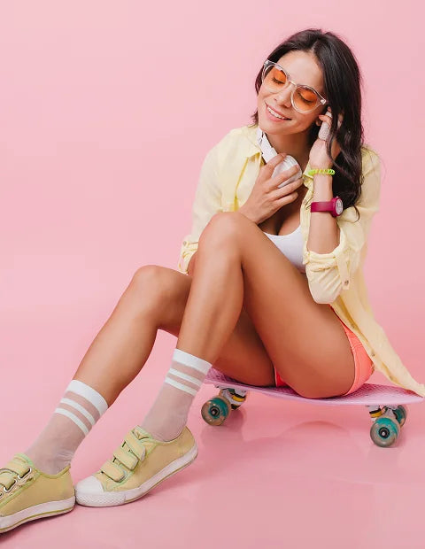 Woman sitting on a skateboard wearing a yellow shirt, pink shorts, and beige sneakers on a pink background